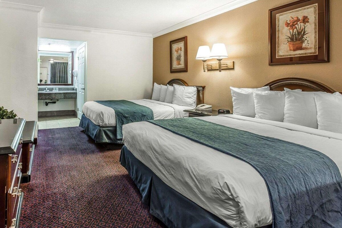 A hotel room with two beds, a desk, a chair, a lamp, and a painting on the wall.