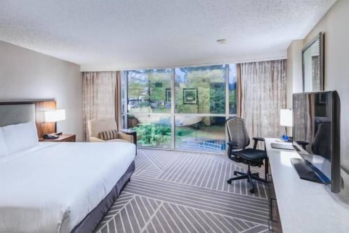Ein Hotelzimmer mit Bett, Schreibtisch mit Stuhl, Fernseher und einem großen Fenster mit Blick auf Grünflächen.