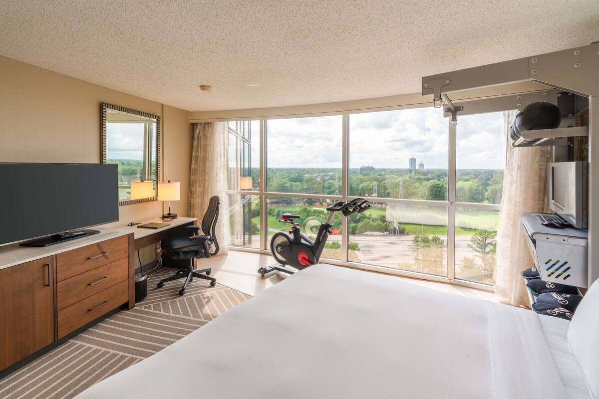 Ein Hotelzimmer mit einem großen Bett, einem Schreibtisch mit Computer, einem Fernseher, einem Heimtrainer und Blick auf eine Stadtlandschaft durch große Fenster.