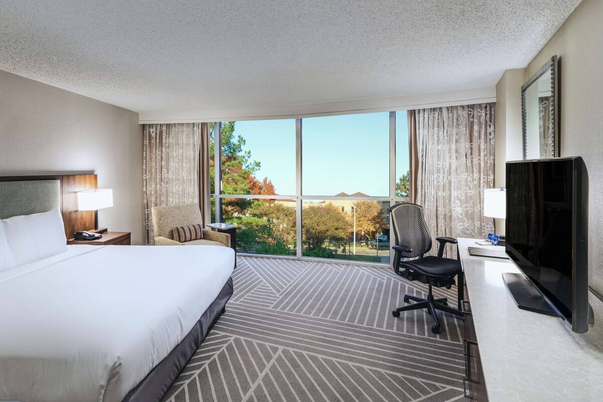 Ein Hotelzimmer mit einem großen Bett, einem Schreibtisch mit Stuhl, einem Fernseher und einem Fenster mit Vorhängen.