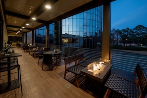Un área de comedor al aire libre con mesas y sillas, una chimenea y vistas al agua y edificios.