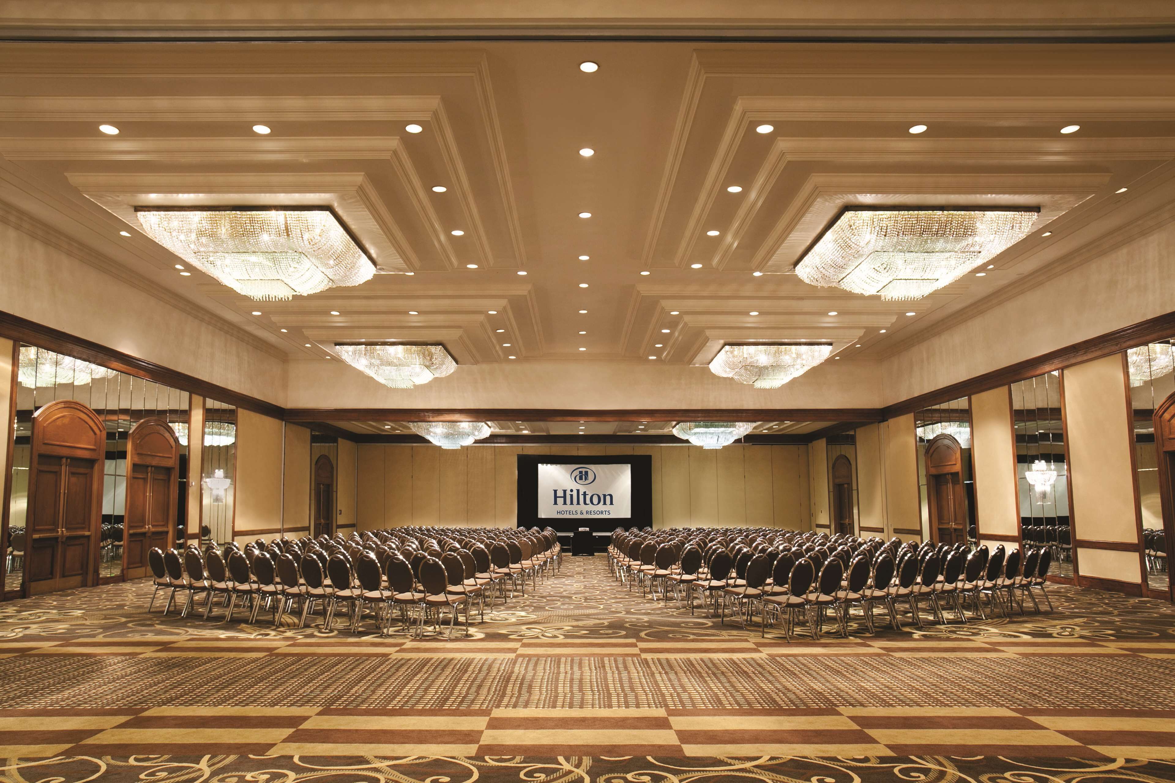 A large conference room with rows of chairs, a stage with a screen displaying 'Hilton Hotels & Resorts', and ornate ceiling lights.