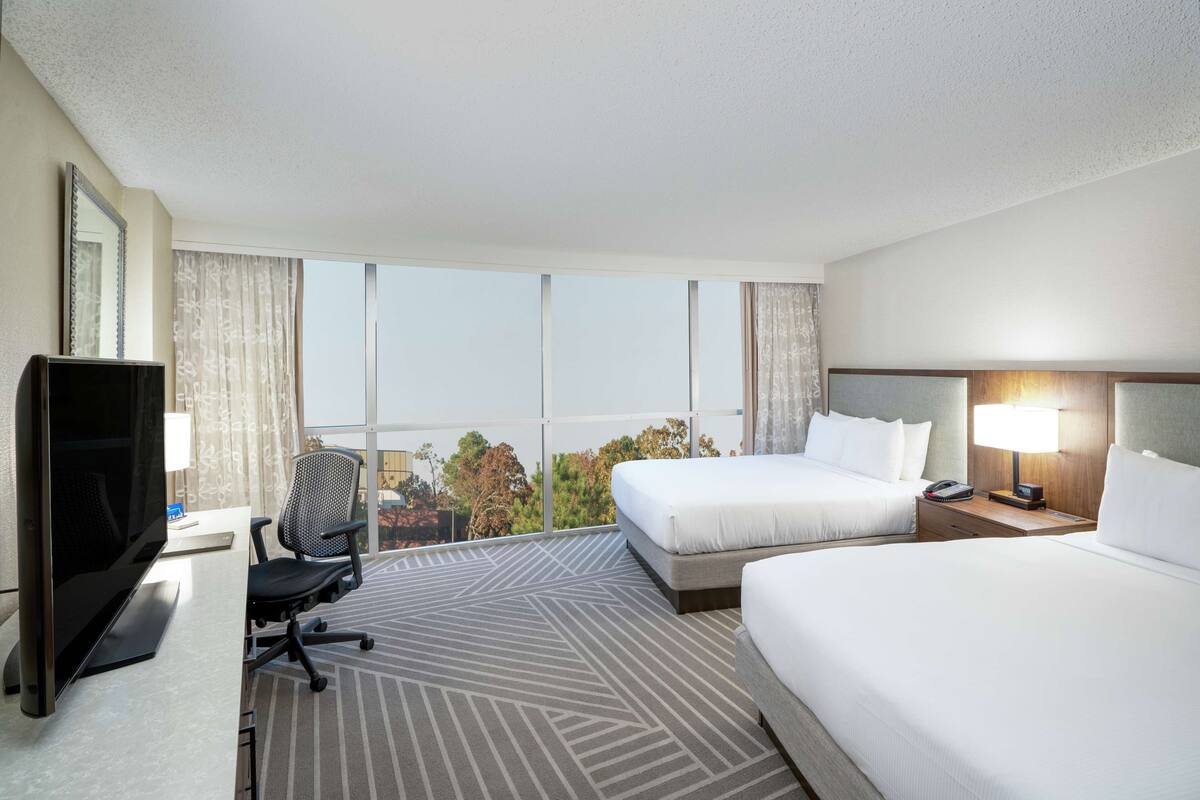 Ein Hotelzimmer mit zwei Betten, einem Schreibtisch mit Fernseher, einem Sessel und einem großen Fenster mit Blick auf die Stadt.