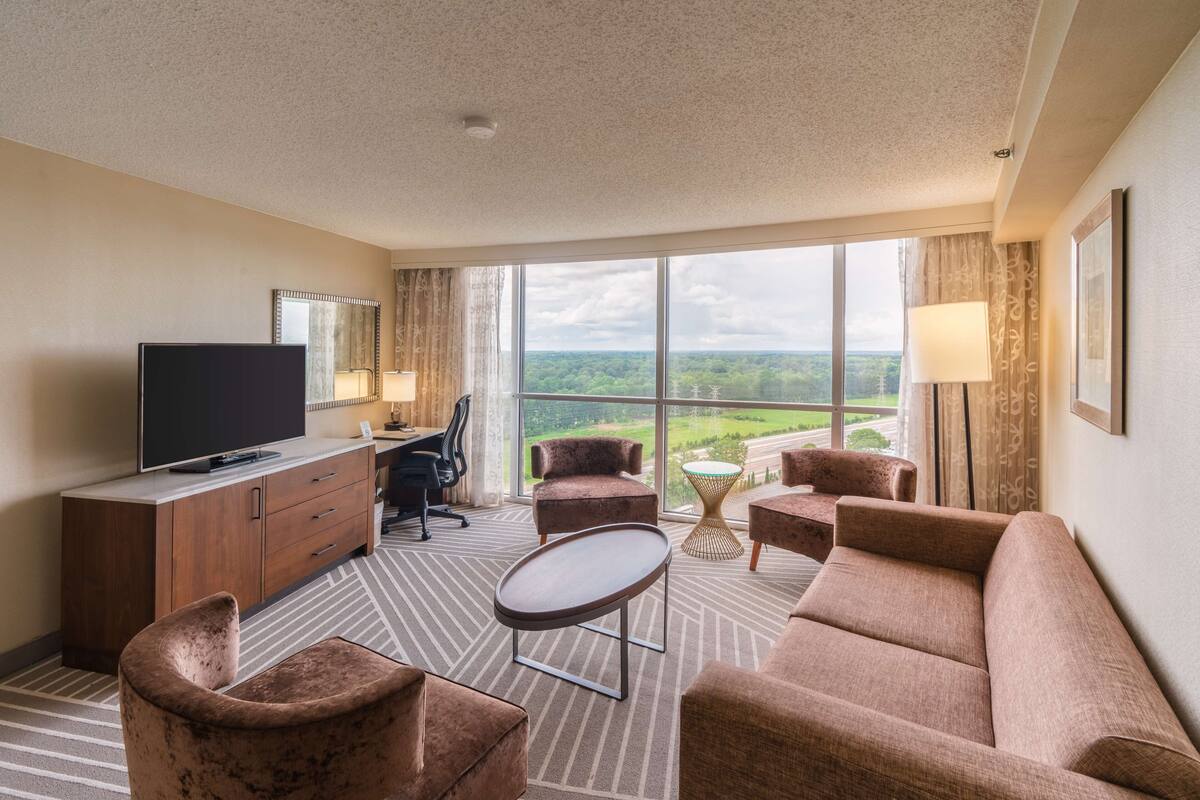 Ein Hotelzimmer mit großem Fenster, Flachbildfernseher, Sofa, Sessel, kleinem Tisch und Schreibtisch mit Stuhl.