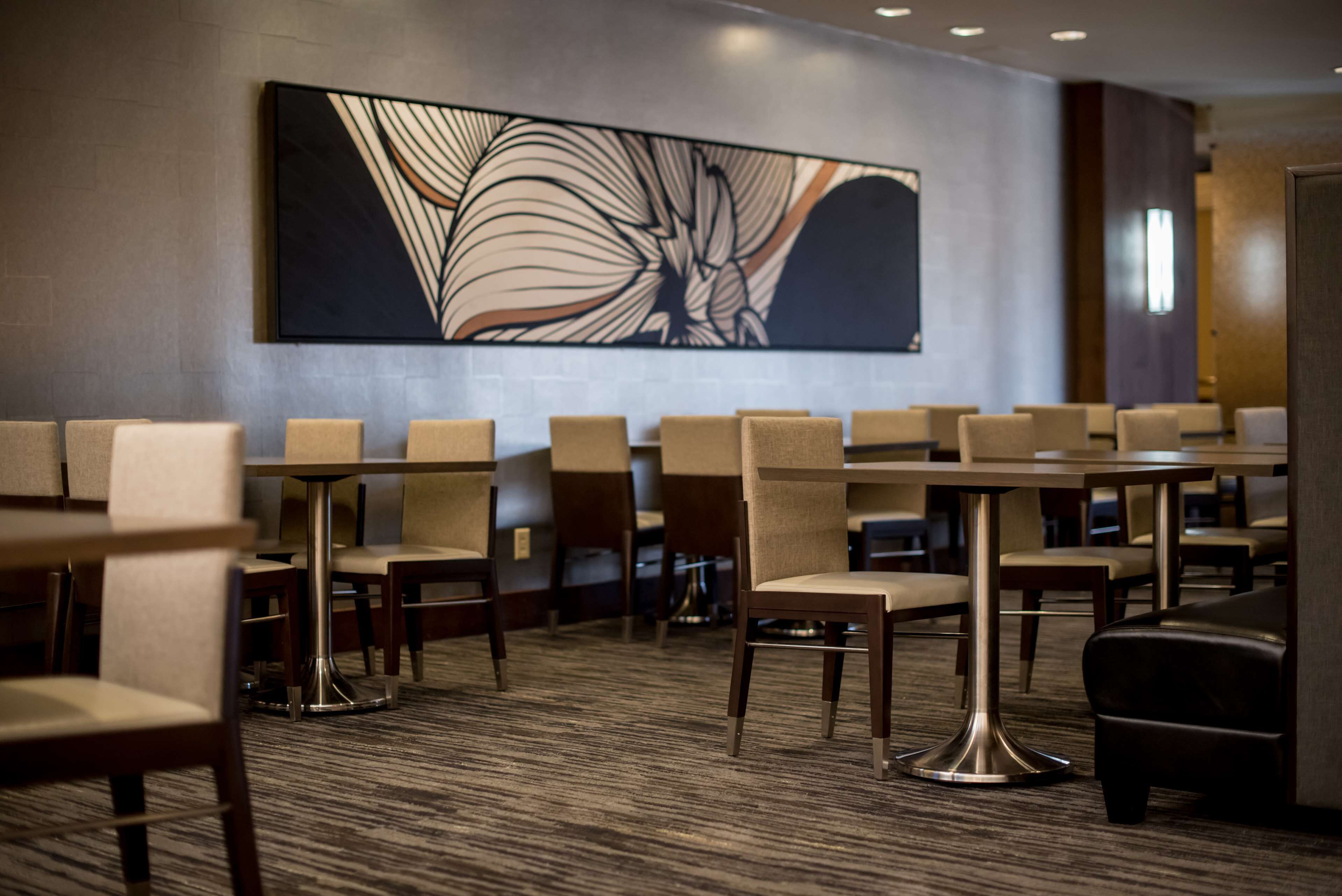 A hotel lobby with multiple tables and chairs, a large abstract artwork on the wall, and a carpeted floor.