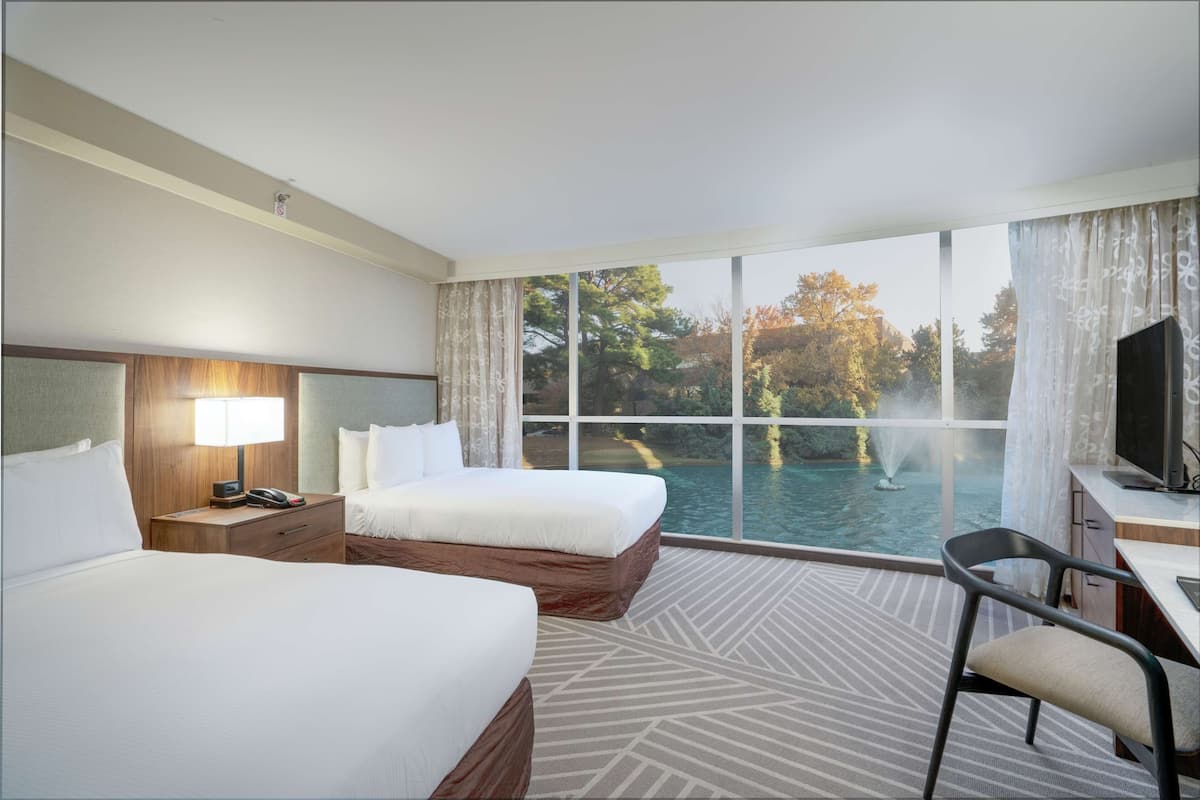 Ein Hotelzimmer mit zwei Betten, einem Schreibtisch, einem Stuhl und einem großen Fenster mit Blick auf einen Swimmingpool und Bäume.