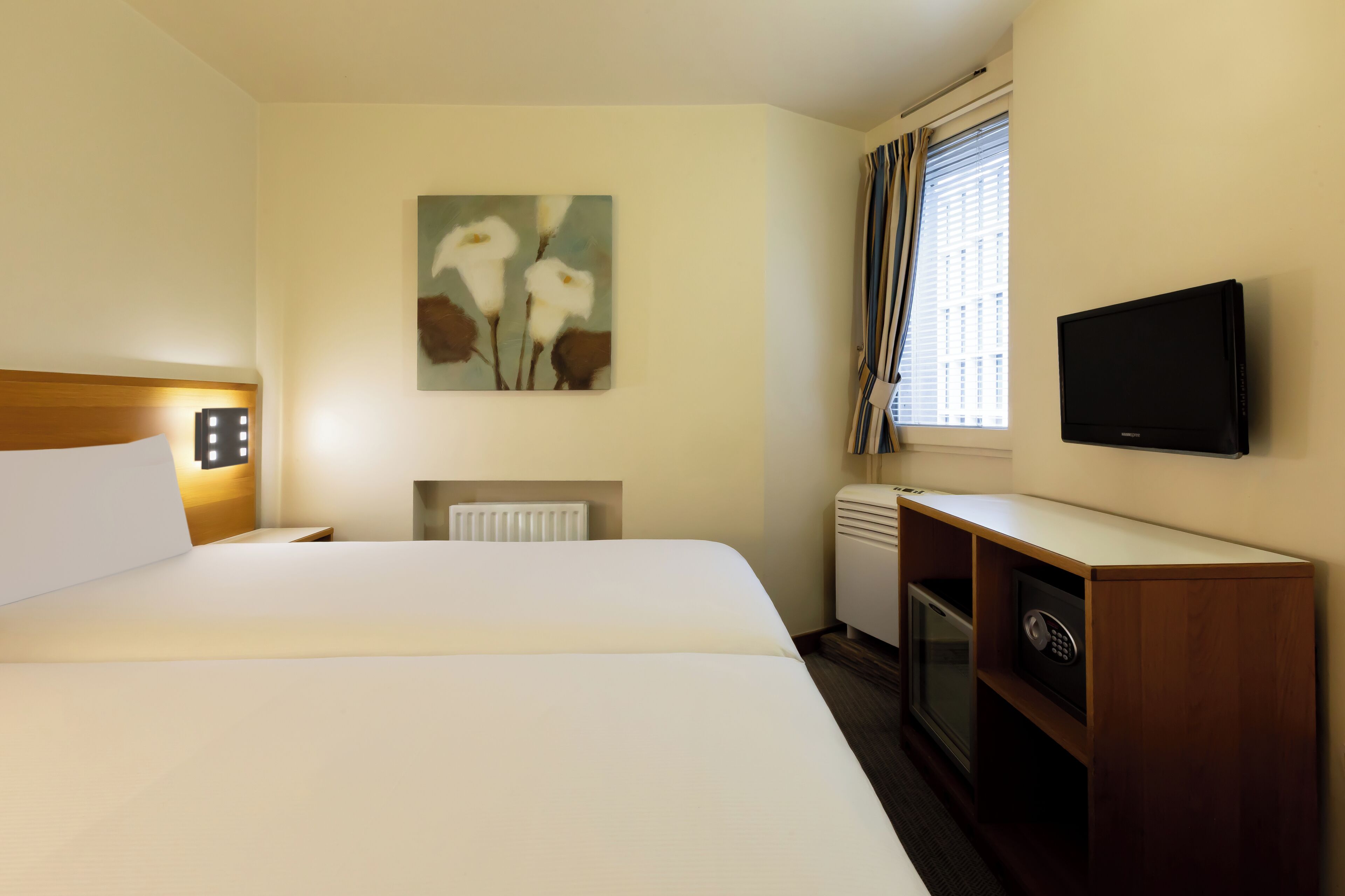 Habitación de hotel con cama, un cuadro en la pared, televisor, minibar y ventana con cortinas.