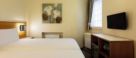 A hotel room with a bed, a painting on the wall, a television, a minibar, and a window with curtains.