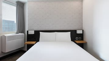 Hotellihuone, jossa on suuri sänky, ikkuna, jossa on sälekaihtimet, geometrisesti kuvioitu seinä ja yöpöytä, jossa on lamppu.