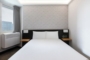 Camera d'albergo con un letto grande, una finestra con persiane, una parete con motivo geometrico e un comodino con una lampada.