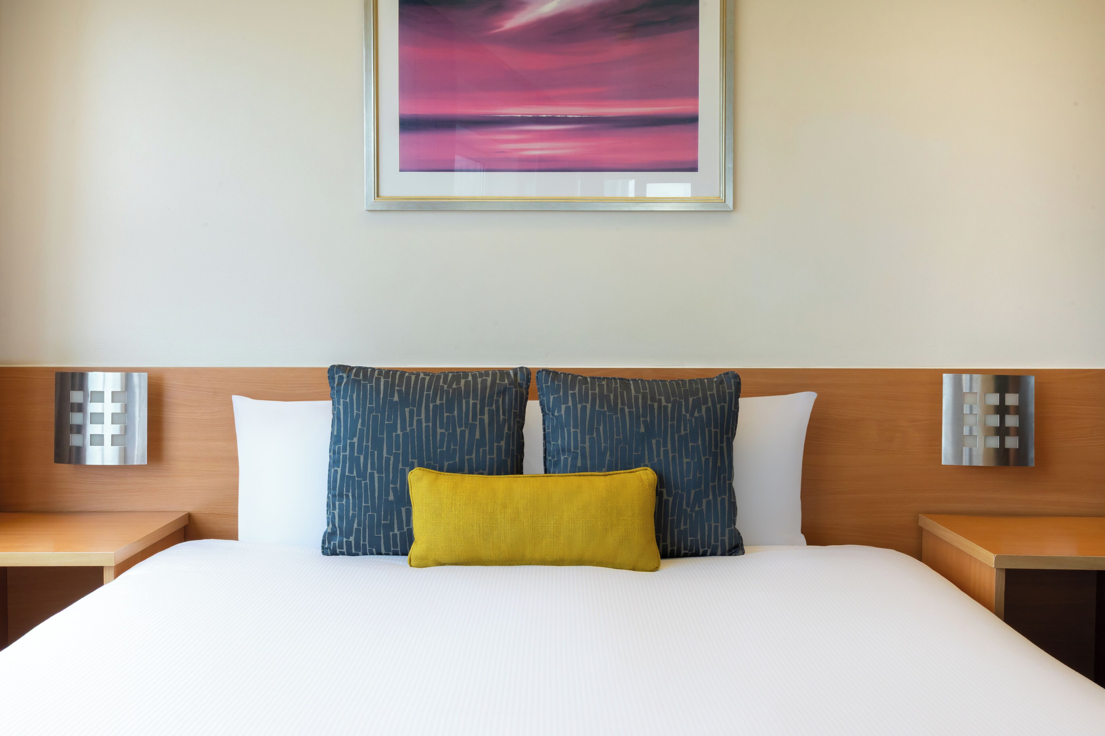A hotel room with a bed, two pillows, a nightstand, and a wall-mounted light.