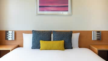 A hotel room with a bed, two pillows, a nightstand, and a wall-mounted light.