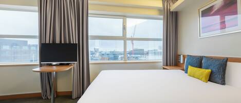 A hotel room with a large bed, a TV on a small table, a window with curtains, and a city view.