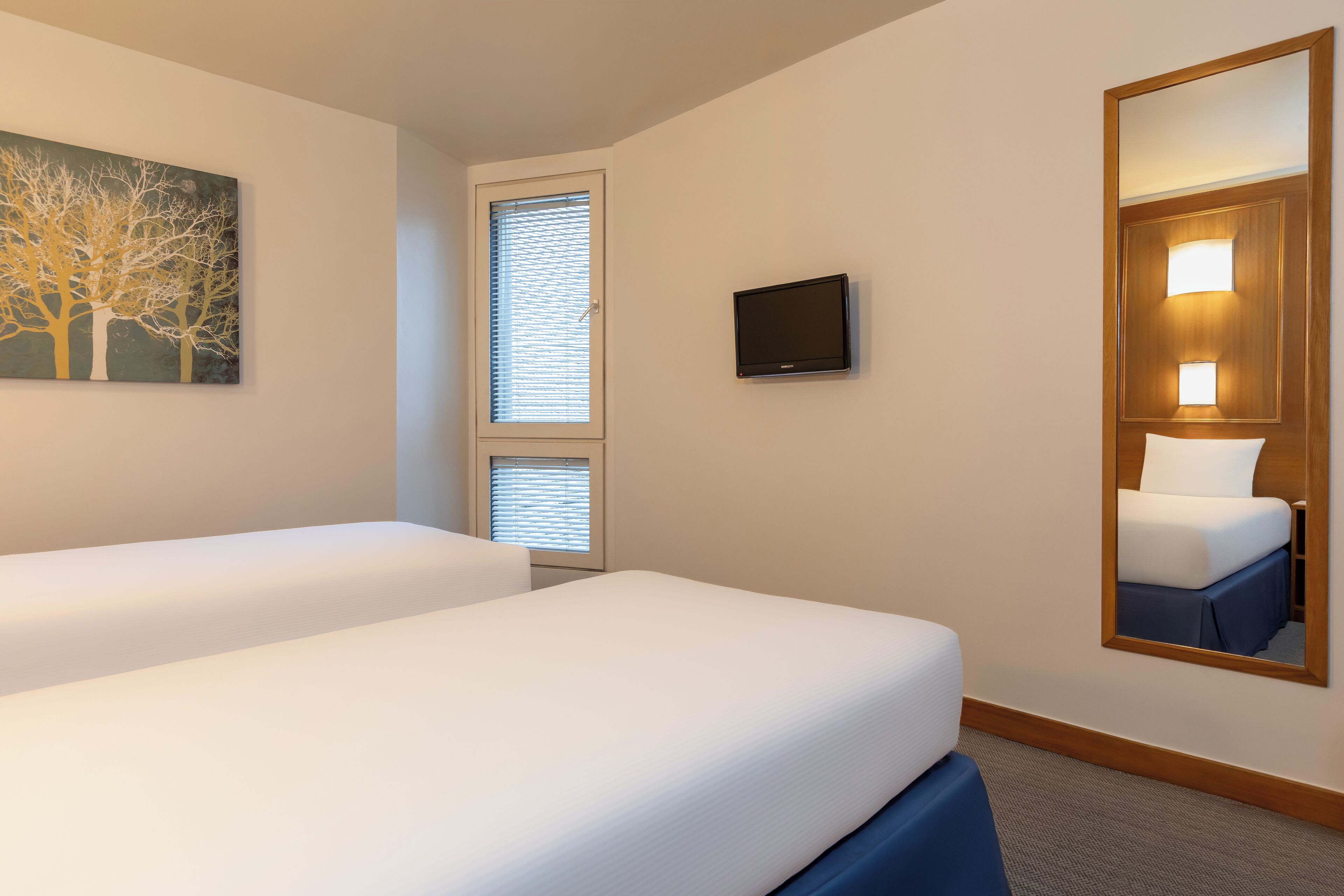 Ein Hotelzimmer mit zwei Betten, einem Fernseher, einem Gemälde und einem großen Spiegel.