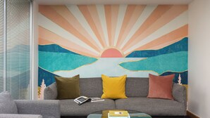 A modern living room with a grey sofa, two cushions, a glass coffee table, and a large wall mural depicting a sunrise over a landscape. -  STG Hotel London Oxford Street (London)