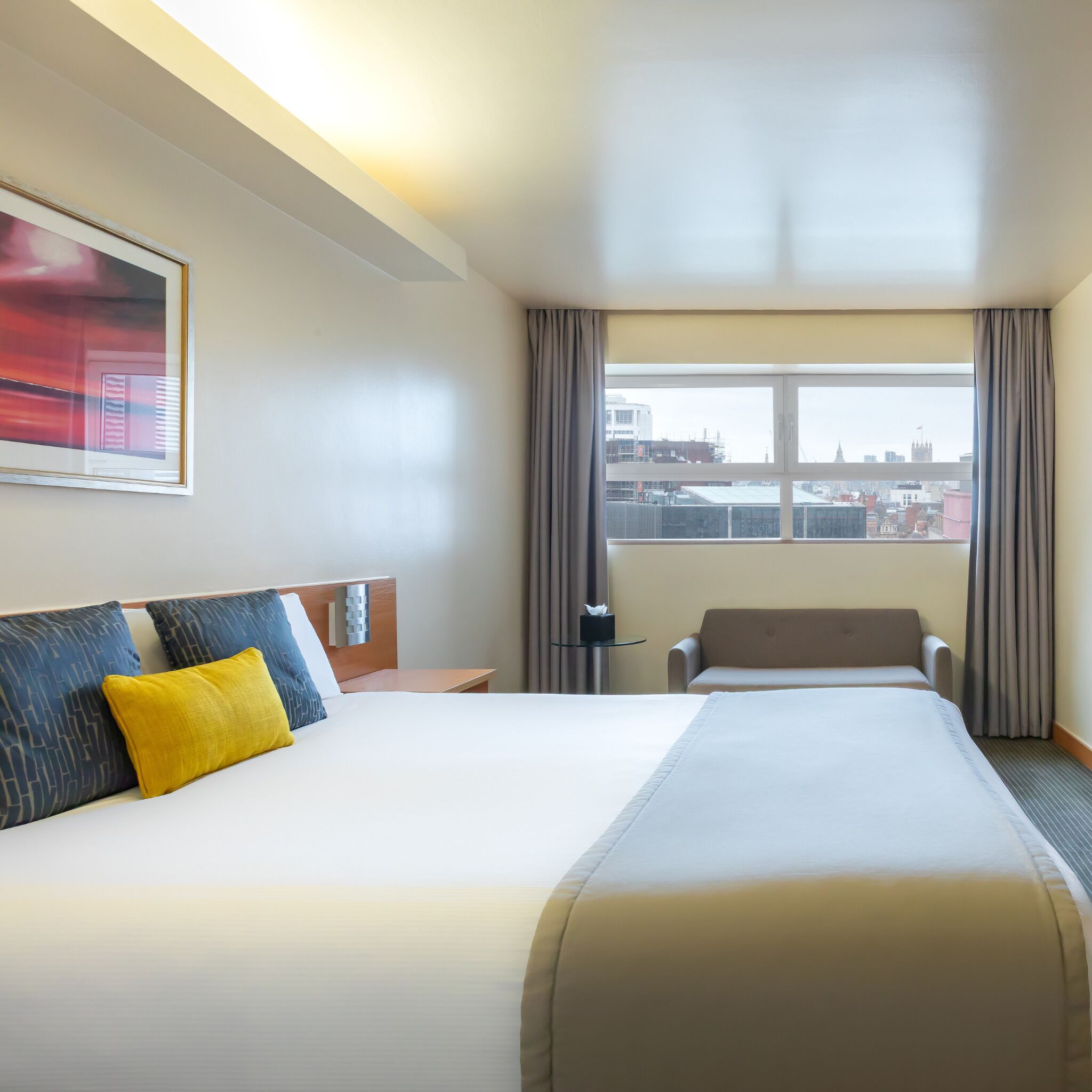 A hotel room with a large bed, a desk, a chair, a window with curtains, and a city view.