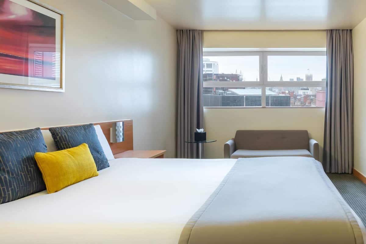 A hotel room with a large bed, a desk, a chair, a window with curtains, and a city view.