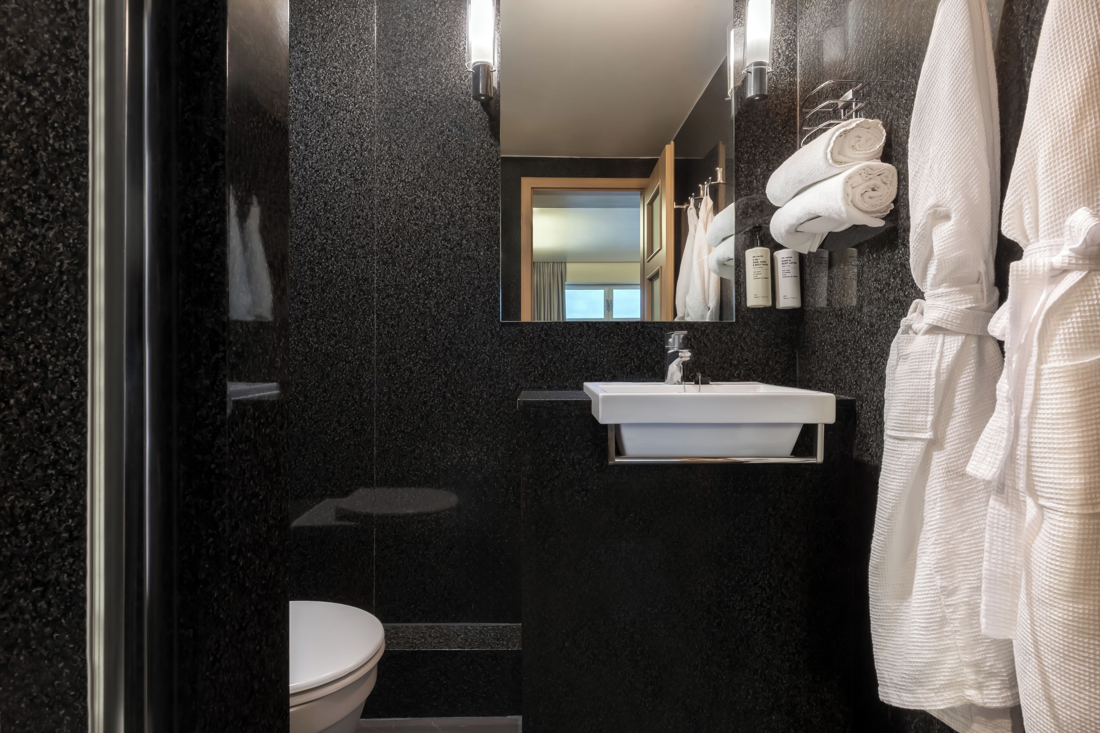A modern bathroom with a black marble wall, a white sink, a mirror, and a white bathrobe hanging on the wall.