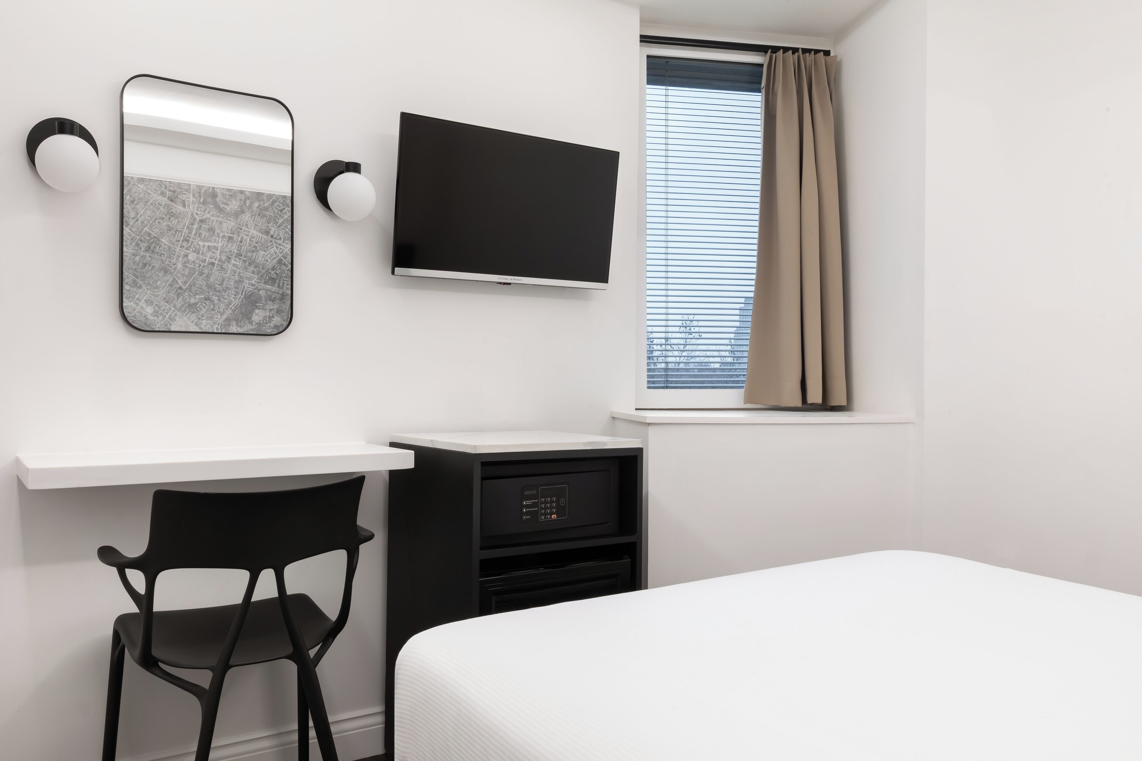Une chambre d’hôtel moderne équipée d’un lit, d’un bureau avec une chaise, d’un téléviseur à écran plat, d’un miroir et d’une fenêtre avec des rideaux.