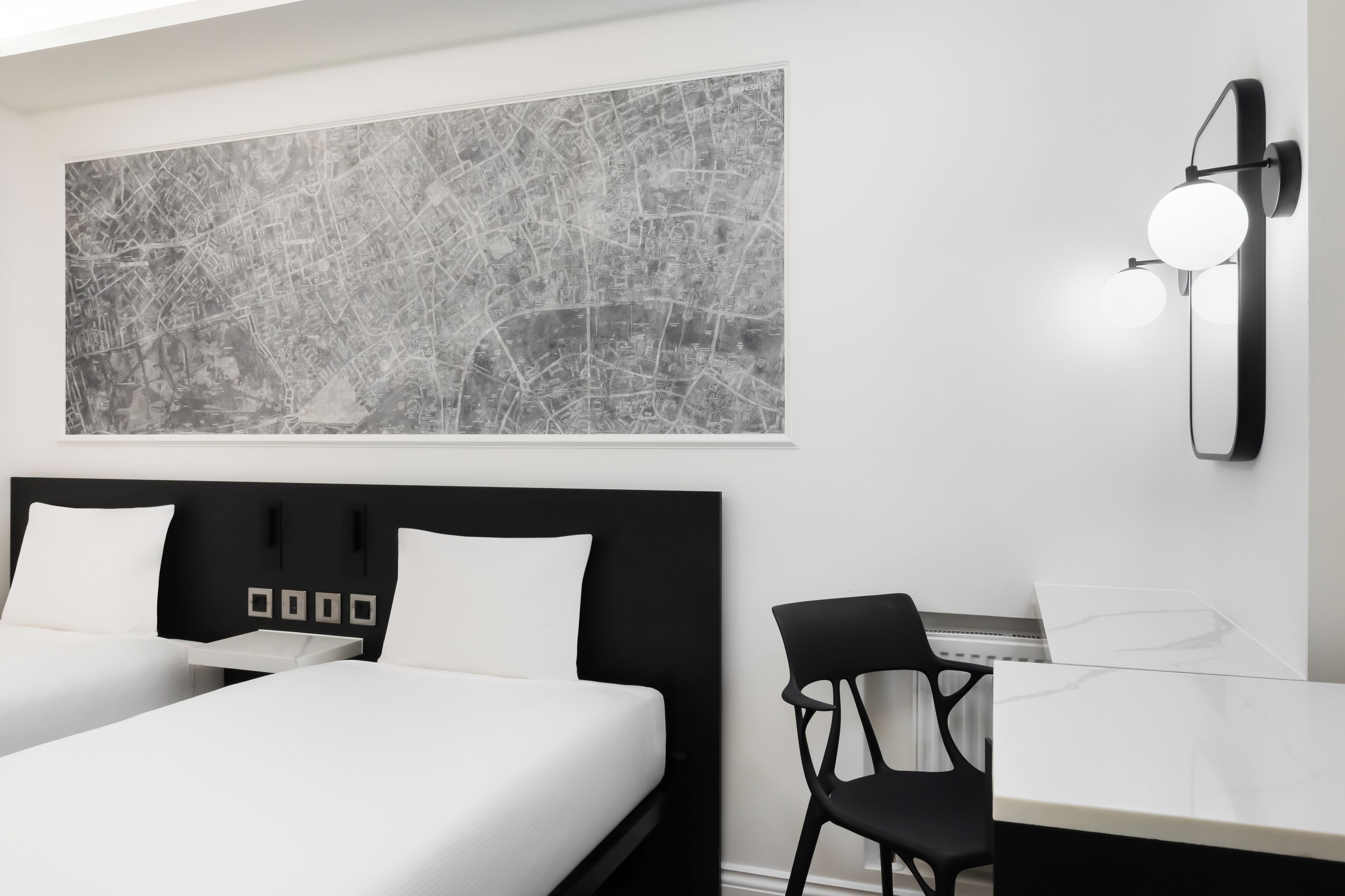 Een moderne hotelkamer met een groot abstract kunstwerk van een stadsgezicht, een zwart hoofdbord, twee bedden, een bureau met stoel en een wandlamp.