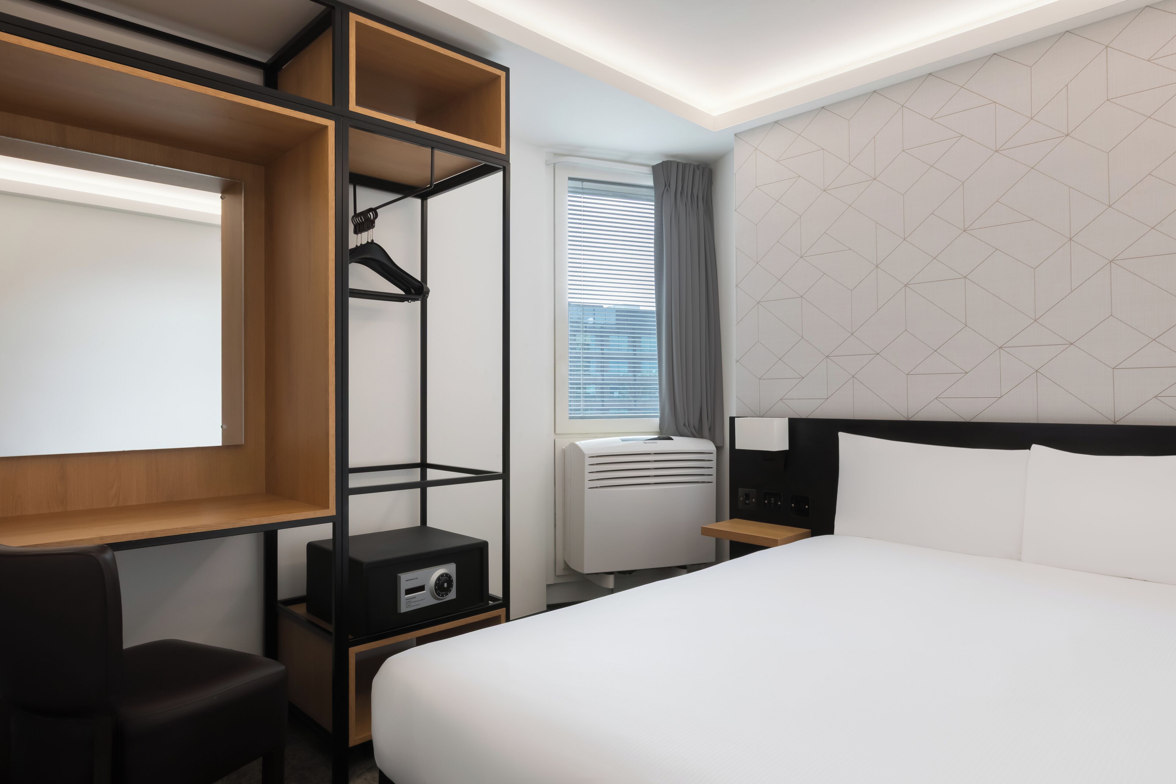 A modern hotel room with a large bed, a desk with a chair, a wardrobe, a radiator, and a window with blinds.