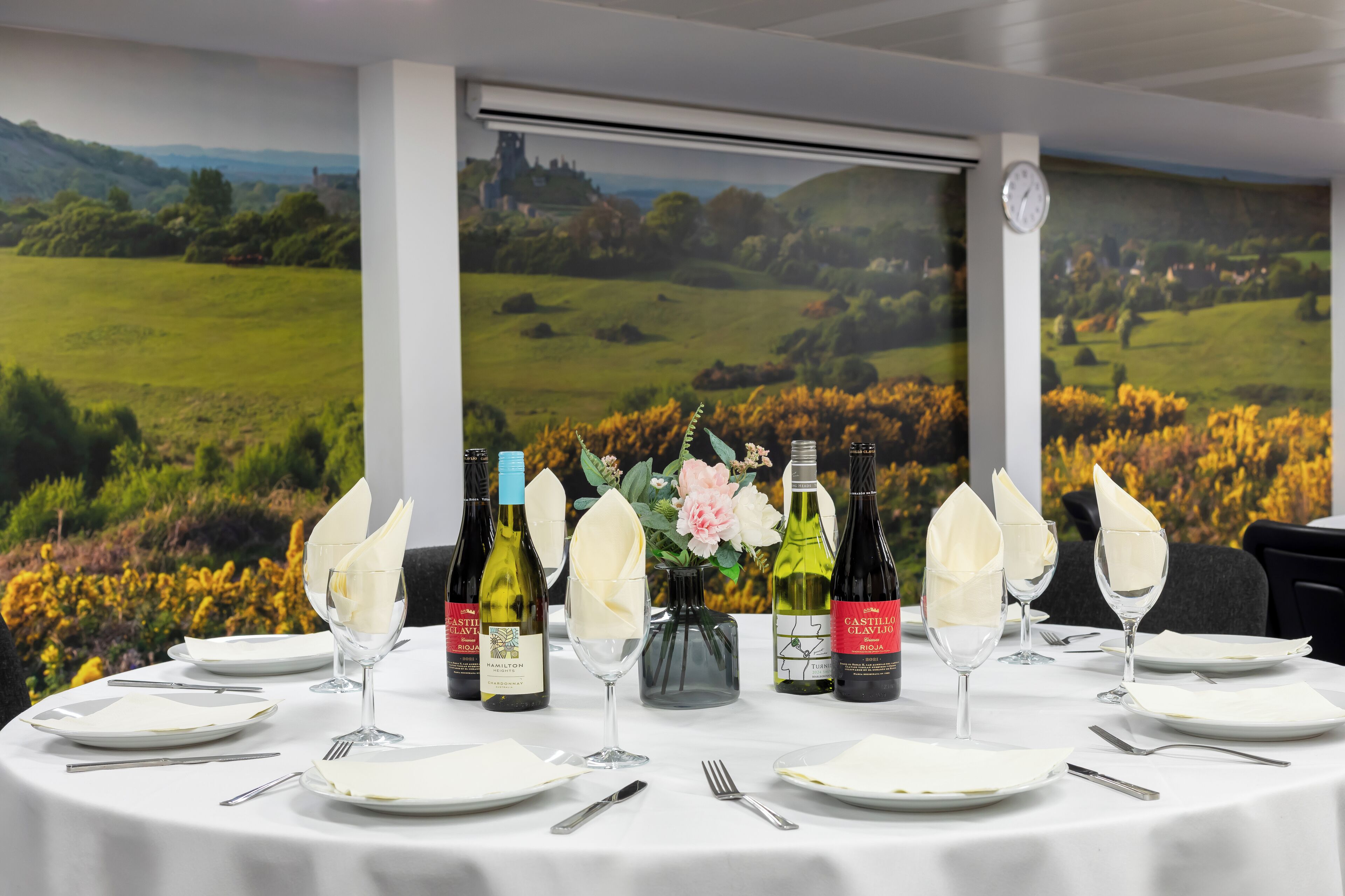 Una mesa de comedor dispuesta con platos blancos, vasos y cubiertos, con botellas de vino y un florero con flores.
