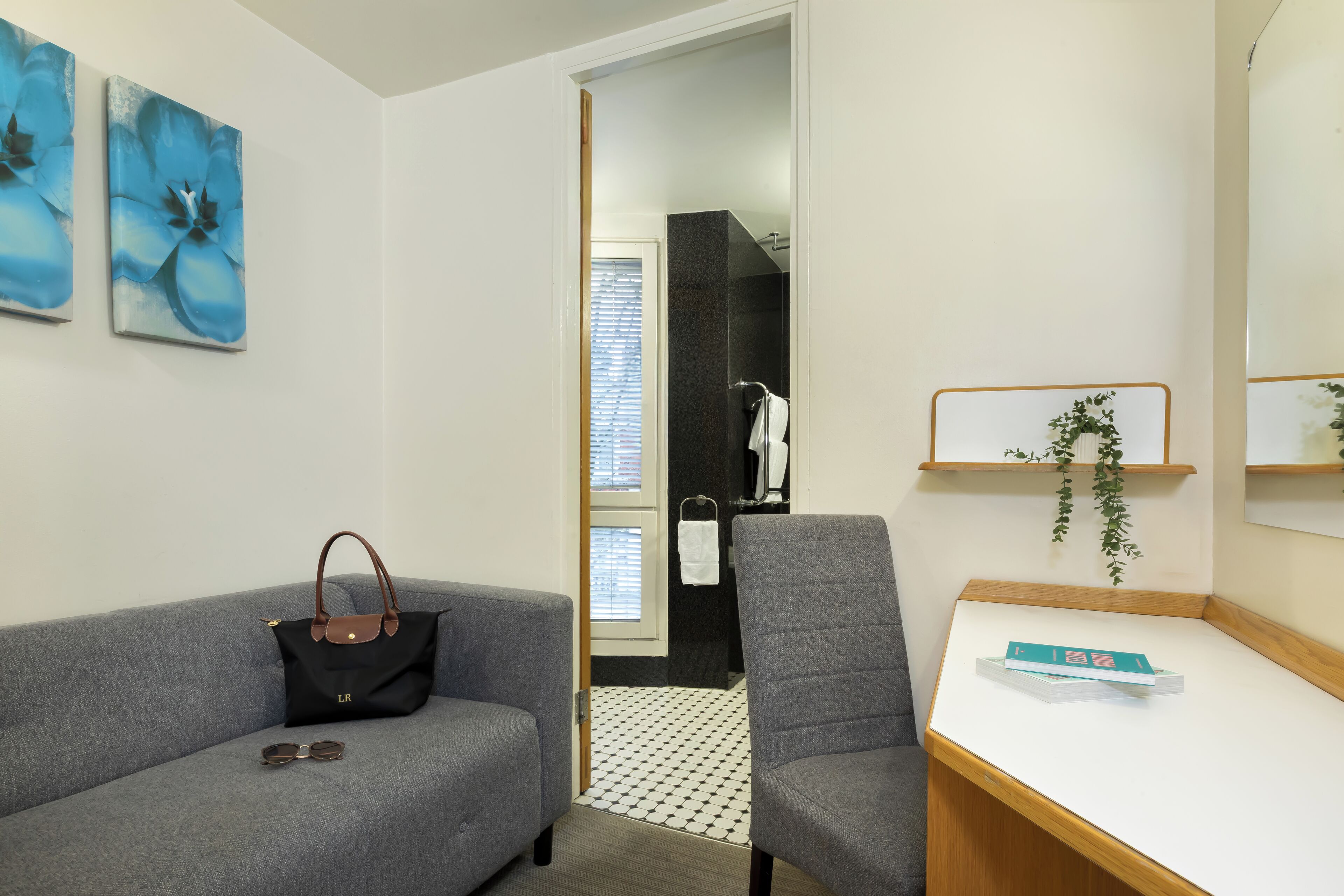 A modern hotel room with a grey sofa, a small desk, and a bathroom visible through an open door.