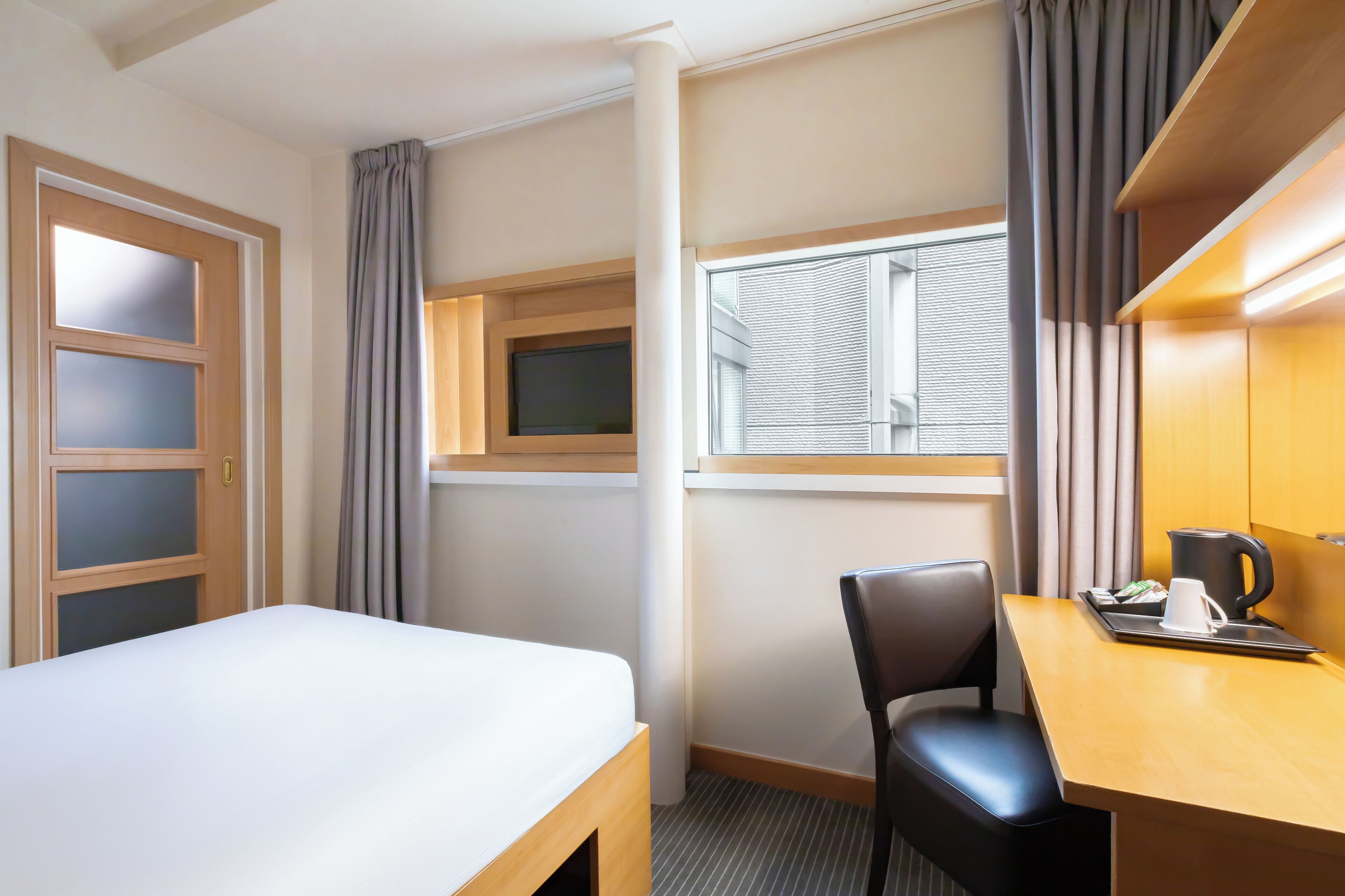 A hotel room with a bed, a desk with a chair, a coffee maker, and a window with blinds.