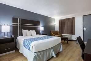 A hotel room with a large bed, bedside tables, a chair, a desk, and an air conditioning unit.