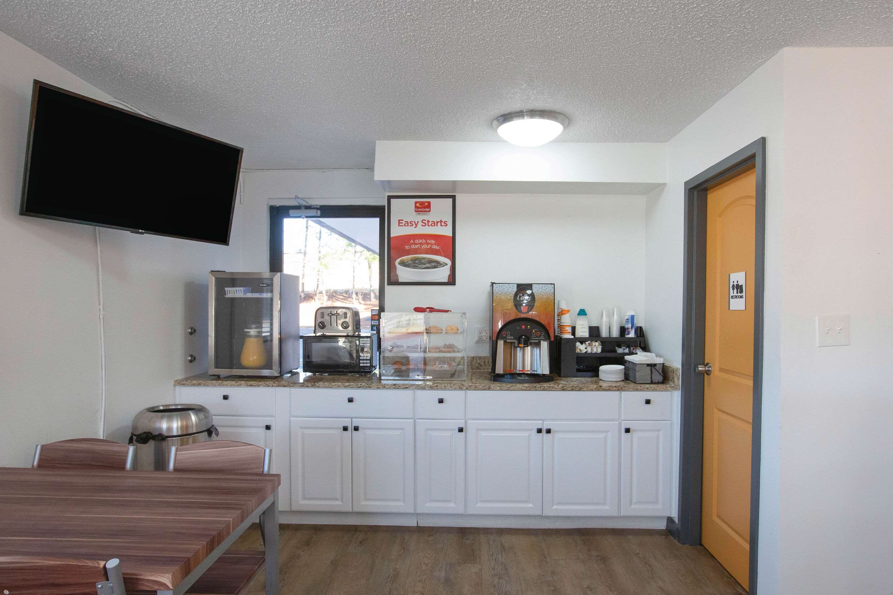 A compact kitchen with white cabinets, a microwave, a coffee machine, and a toaster. There's a TV mounted on the wall and a door to the right.