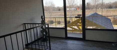A room with a wooden floor, a black railing, and a large window offering a view of a building with a blue roof and a yellow staircase.