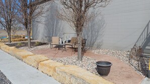 En udendørs patio med betongulv, omgivet af en lav stenvæg. To stole og et bord, samt en stor plantekasse i midten.