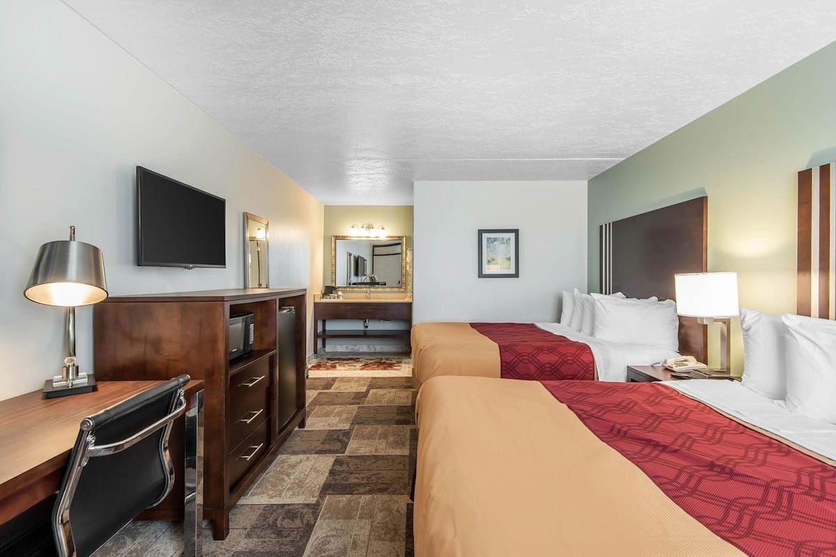 Una habitación de hotel con escritorio, dos camas, televisión, espejo y baño.