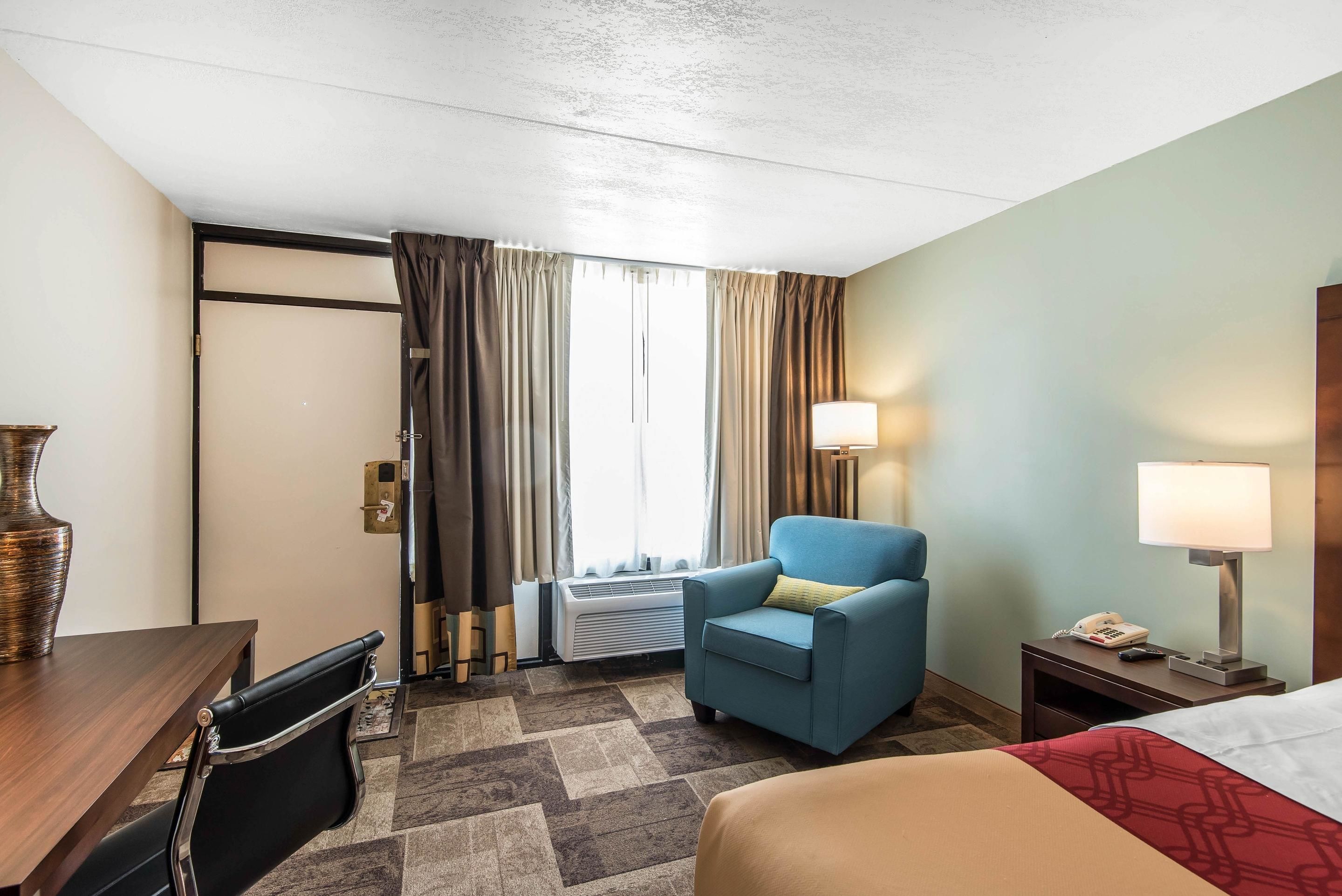 Une chambre d’hôtel avec un lit, un bureau et une chaise, un fauteuil bleu, une table de chevet avec une lampe, et une fenêtre avec des rideaux.