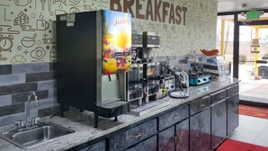 Et morgenmådsområde med juiceudskænker, kaffemaskiner og en væg udsmykket med ikoner af mad og drikke.