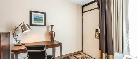 A hotel room with a wooden desk, a black office chair, a lamp, a vase, a framed picture, a door with curtains, and a rug.