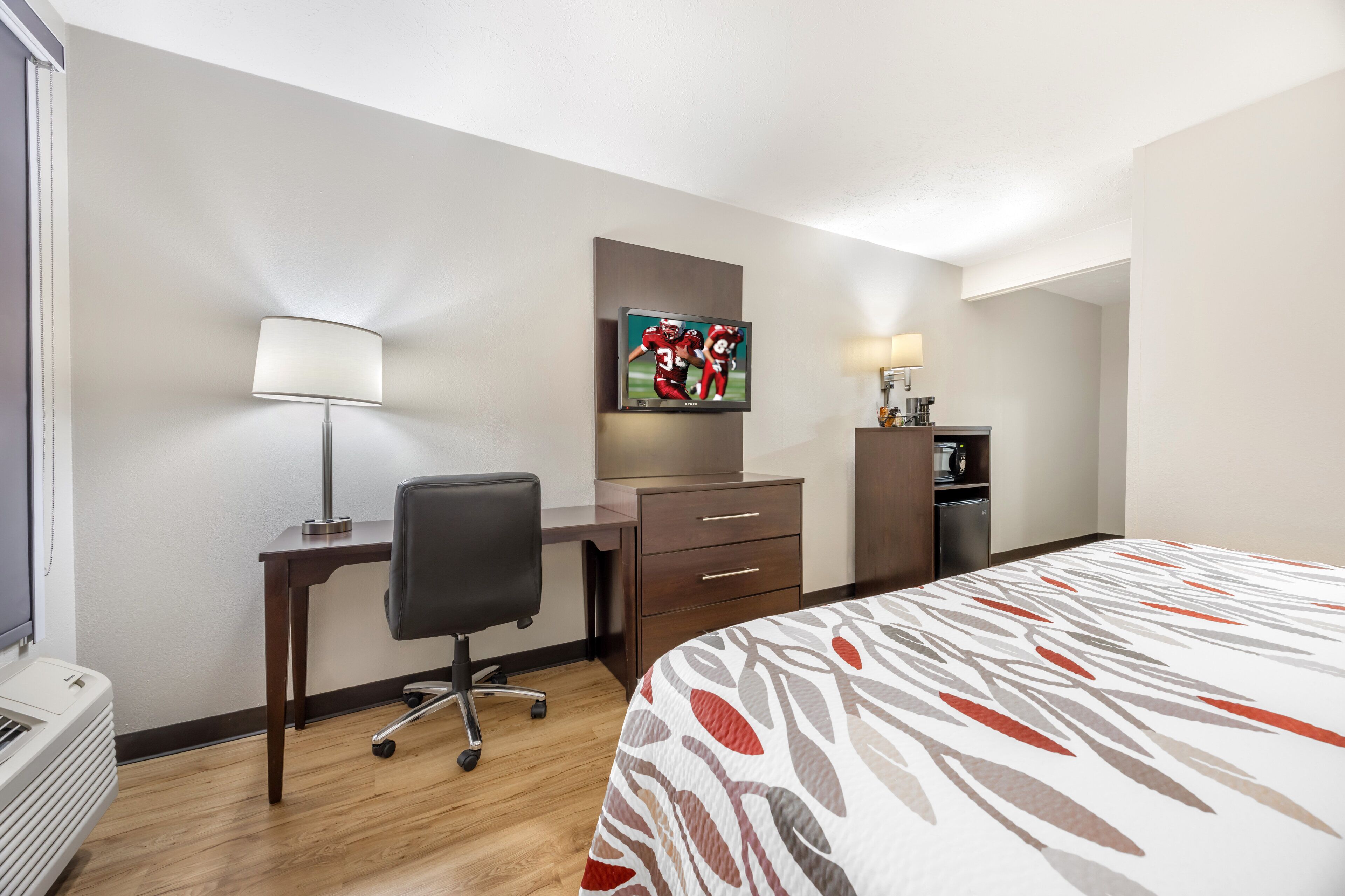 A hotel room with a bed, desk, chair, dresser, and a flat-screen TV mounted on the wall.