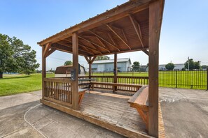 Um gazebo de madeira com bancos e uma mesa, rodeado por um caminho de concreto e um campo verde.
