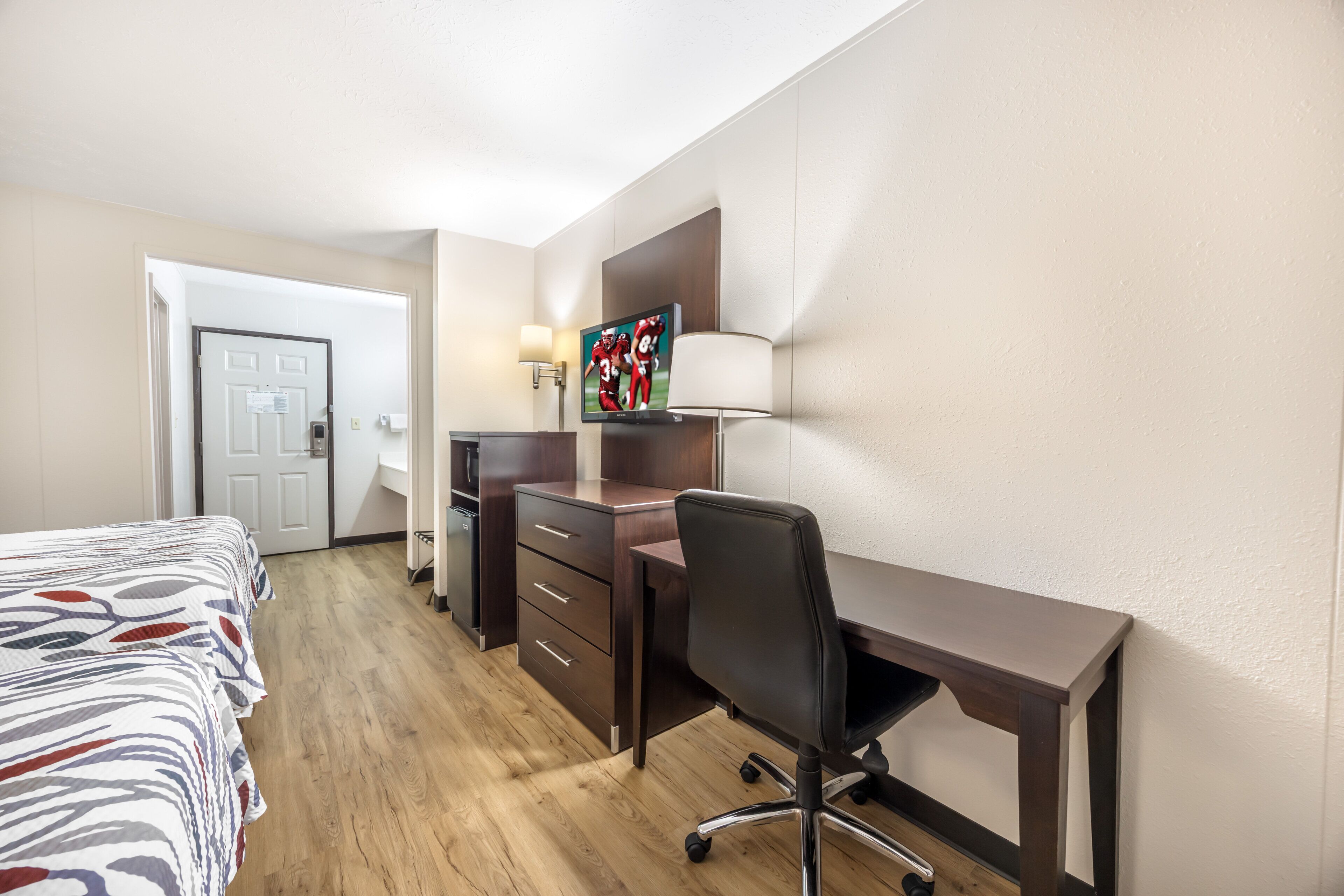 A hotel room with a bed, desk, chair, lamp, TV, and a door.