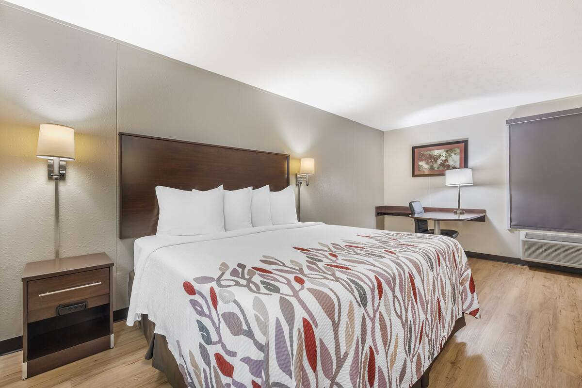Habitación de hotel con una cama grande, dos mesitas de noche con lámparas, un pequeño escritorio con silla y un ventanal amplio.