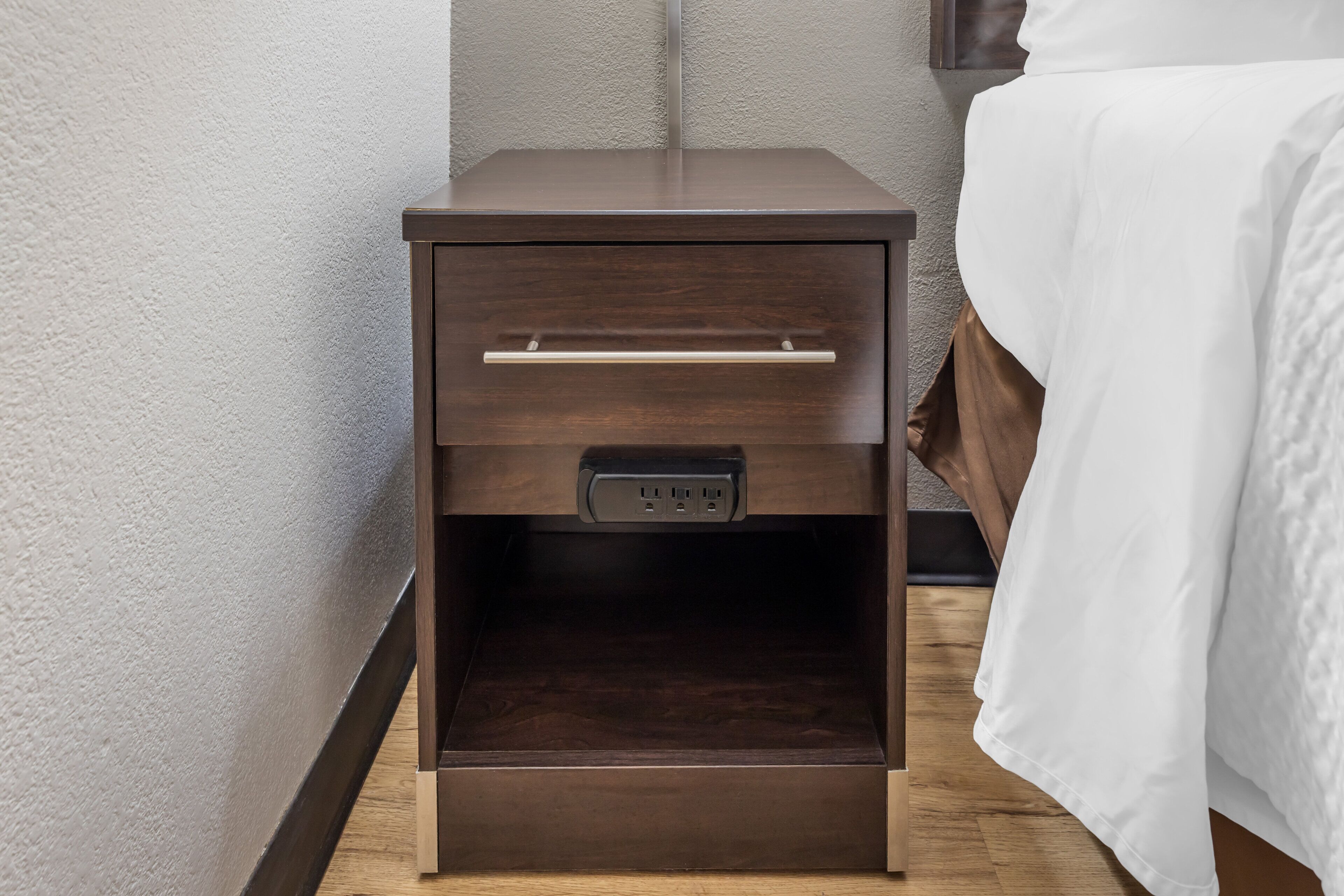 A wooden bedside table with a drawer, a power outlet, and a small shelf.