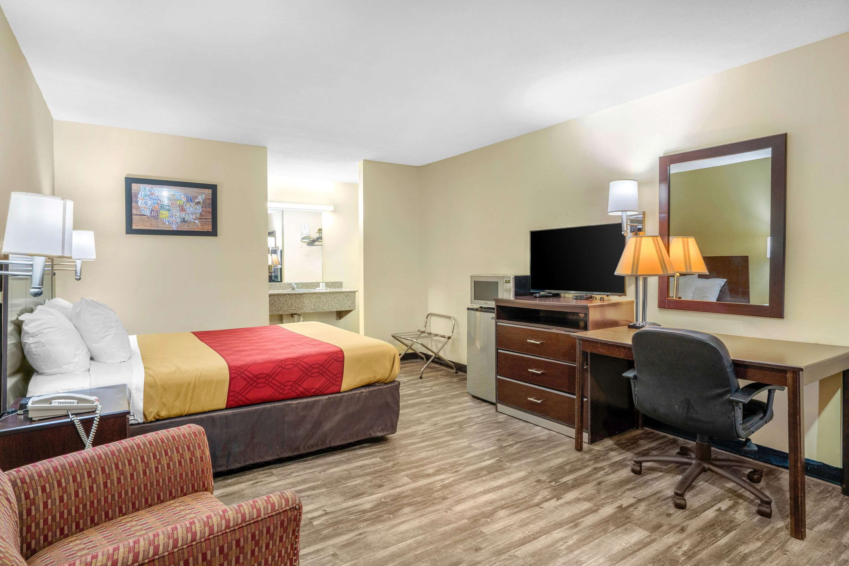 Une chambre d’hôtel comprenant un lit, un bureau avec un ordinateur, une chaise, une lampe de bureau, une télévision et une petite table avec une lampe.