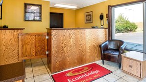 Un área de recepción con un mostrador de madera, un sillón de cuero marrón, una alfombra roja con el logotipo y un ventanal con vistas a zonas verdes.