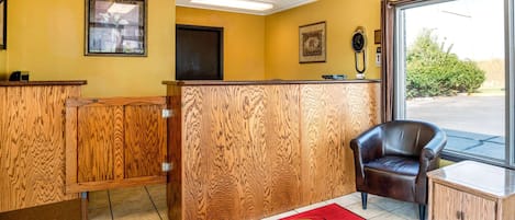 A reception area with a wooden reception desk, a brown leather chair, a red carpet with the logo, and a window with a view of greenery.