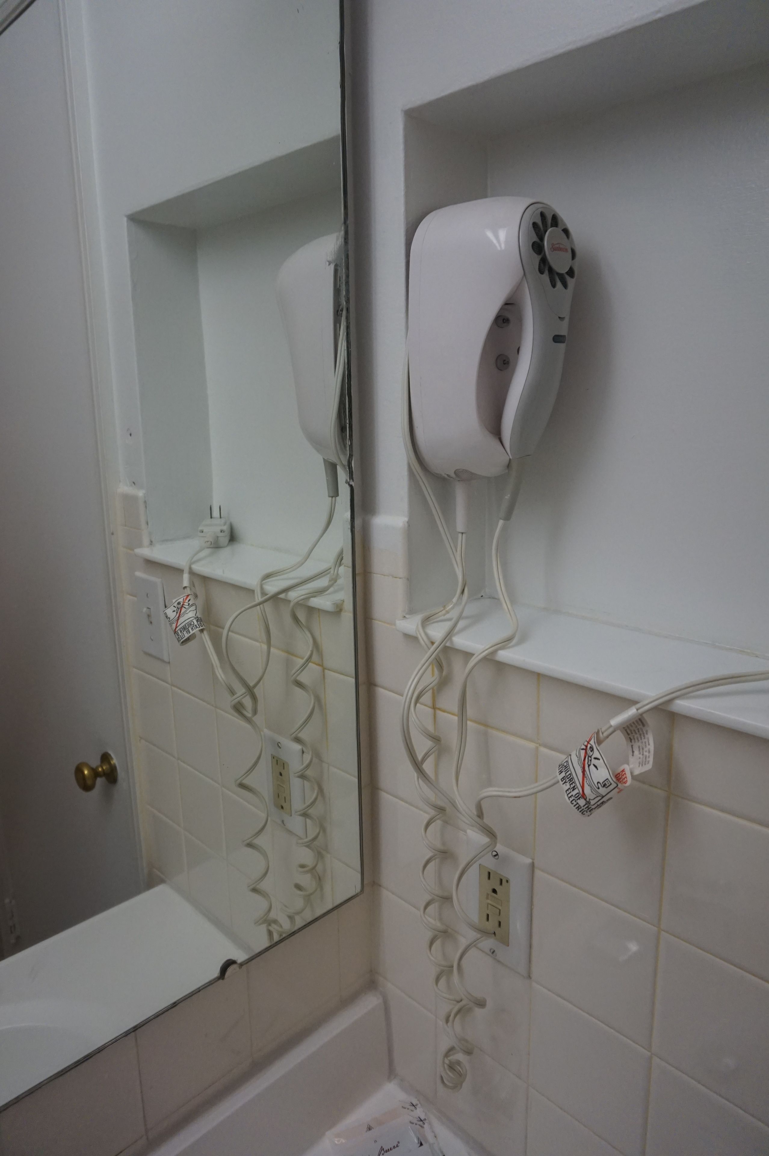 A bathroom with a white tiled wall, a mirror, a hairdryer mounted on the wall, and a shelf with a power strip.