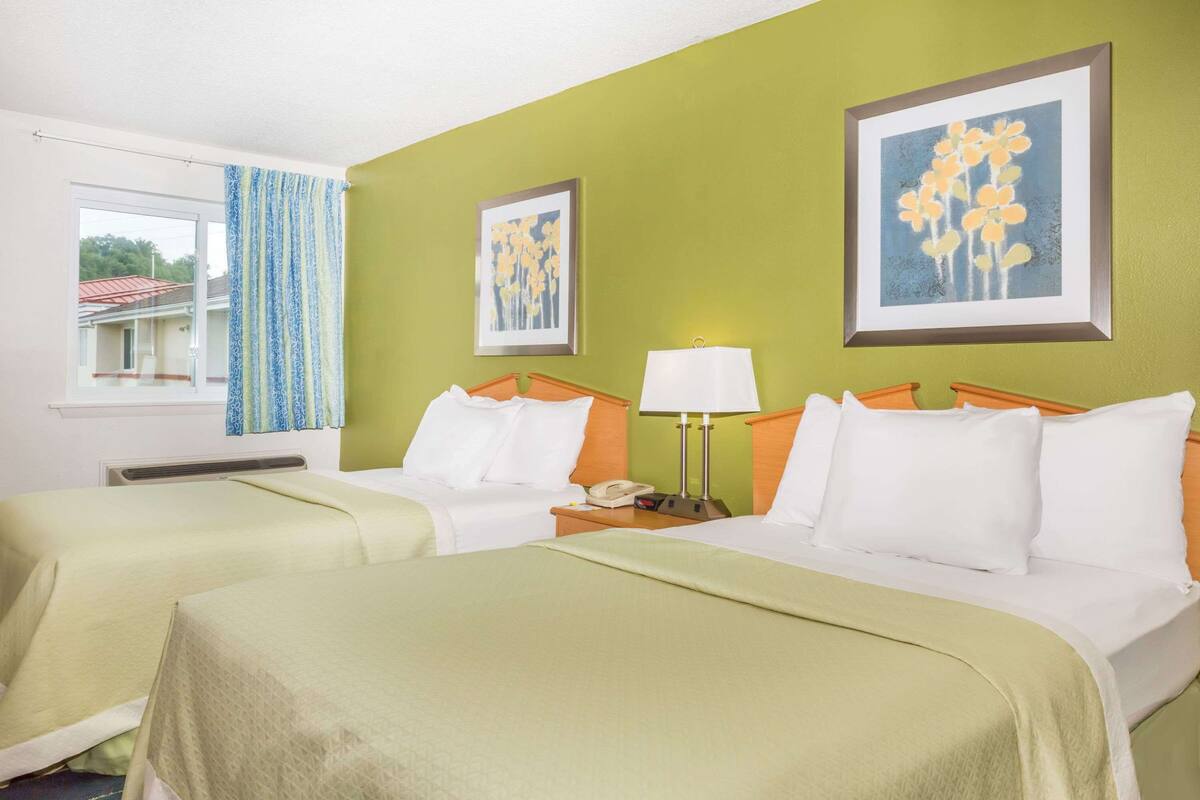 A hotel room with two beds, a window with curtains, a framed picture on the wall, and a bedside lamp.