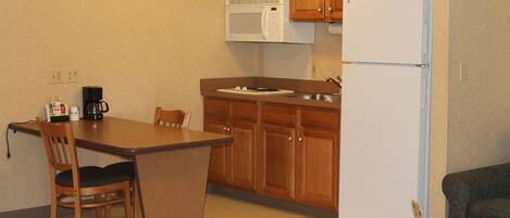 A compact kitchen with wooden cabinets, a white refrigerator, a microwave, a coffee maker, and a small dining area with a chair.