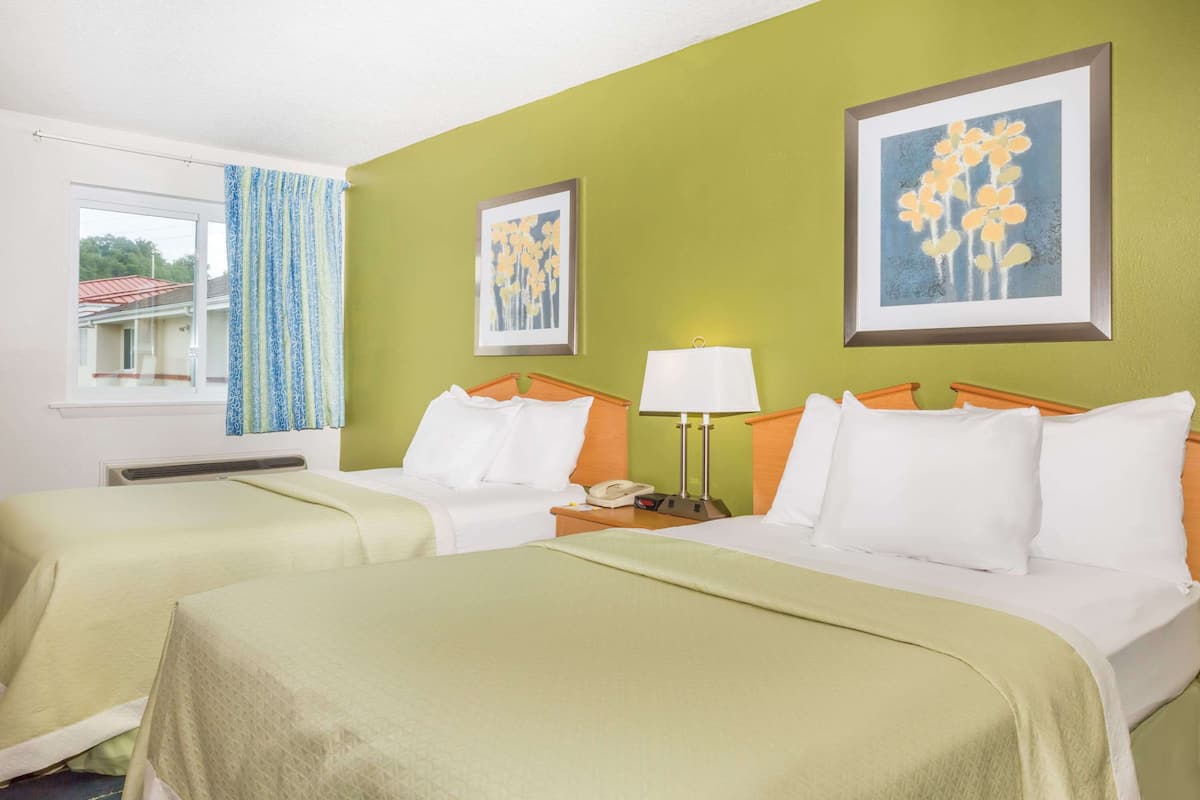 A hotel room with two beds, a window with curtains, a wall with two framed pictures, and a bedside table with a lamp and phone.