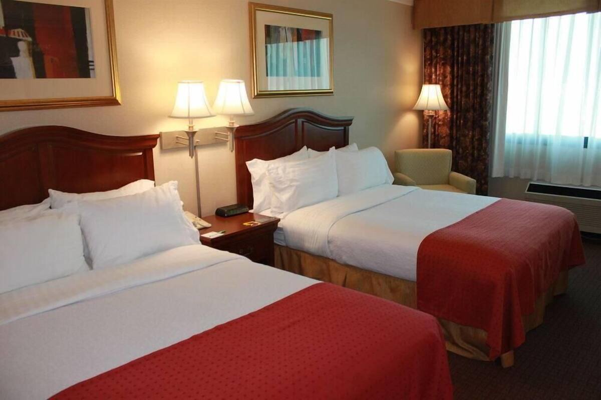 A hotel room with two beds, a nightstand, a chair, a lamp, and a window with curtains.