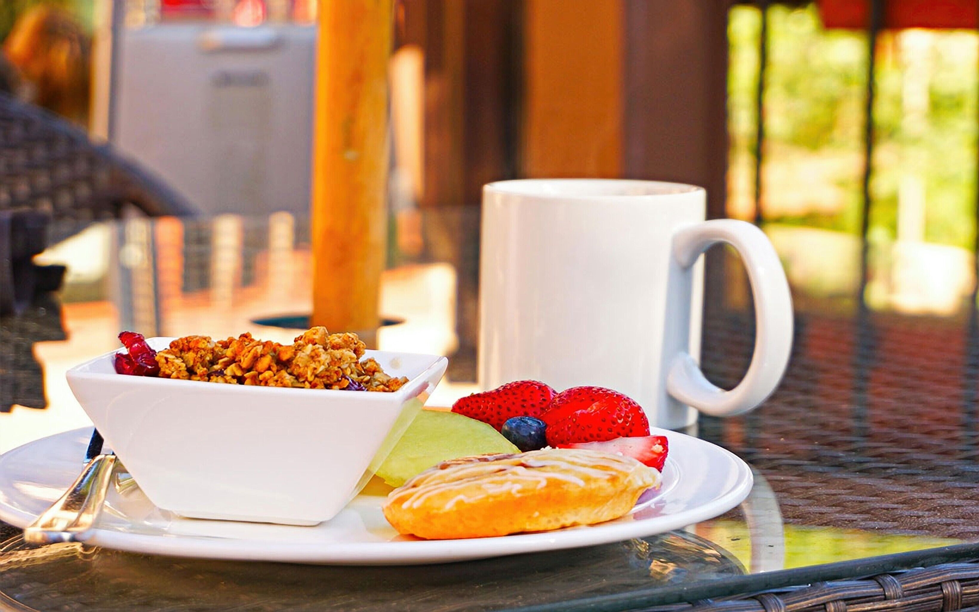 A breakfast setting with a bowl of granola, fresh fruits, and a pastry on a plate, accompanied by a white mug.